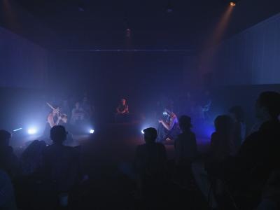 Samita Sinha, Okwui Okpokwasili, and Ash Fure sit in a triangular formation under warm spotlights among a seated crowd in the darkened Grand Luxe Hall. Brilliant blue and white lights positioned near them on the floor illuminate the space with a soft, cool glow.