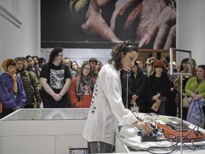 Katayoon Yousefbigloo stands behind a counter covered by guitar pedals connected to a red electric guitar. She looks downwards at a knob she adjusts on one of the guitar pedals. The crowd that fills the gallery space to her side follows her line of sight towards the pedal.