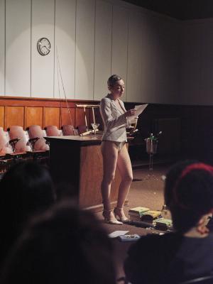 Marie Segolene Brault stands and reads from a paper while spotlit in the Grand Luxe Hall. Piles of books rest at her feet and the heads of an onlooking audience frame her position.