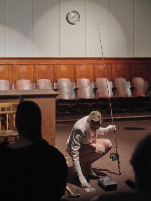 Marie Segolene Brault kneels down with a fishing rod in her right hand. With her left hand, she grabs a cassette resting on the floor next to an open cassette player.