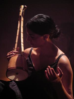 Samita Sinha holds an ektara close to her chest with one hand. Her other hand rests in the air with its palm facing upwards. Warm overhead lighting and hints of orange separate her body from the dull purple hued background as her head turns towards the instrument.