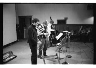 A black-and-white photo of David Prentice playing violin, Bill Smith playing soprano saxophone, and David Lee playing cello. Two audience members can be seen sitting along the wall of the Grand Luxe Hall.