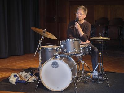 Dylan van der Schyff wears a brown sweater, grey dress pants and pale blue sneakers, as they speak through a microphone, sitting behind a drum set, in the Grand Luxe Hall.