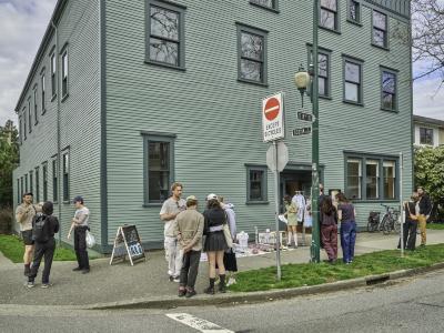 Clusters of people gather on the street in front of Western Front socializing at a sidewalk sale. Clothes are arranged on racks and blankets on the ground.
