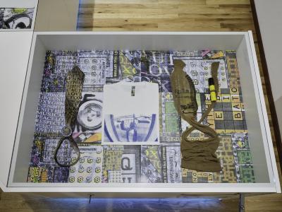 A glass top vitrine displaying a screenprinted neck tie, tee shirt and stockings tied to a pen. The inside of the case is lined with enlarged scans of scratch and win lottery tickets. 