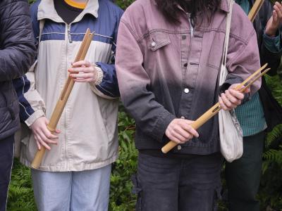 Two people wearing jackets and jeans stand holding bamboo buzzers. Their background is filled with forest greenery and their heads are cut off by the frame.