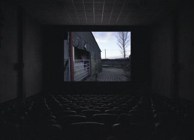 A movie theatre screening shows a rusty metal barn with an opening filled with hay, interrupted by a gate in front of it. A person dressed in a pink jacket and pink rainboots sits on the hay with their legs resting on the gate. A brown dog stares at them from a path to the person’s right, as a brick wall and blue sky sit in the background at the end of the barn.
