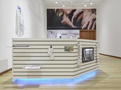 The side of a white slatboard kiosk is adorned with acrylic shelves, a pair of socks, and a television monitor. Icy blue light emanates from below the structure. A billboard sized photograph of a glamorous Chinese grandmother wraps the wall in the background. 