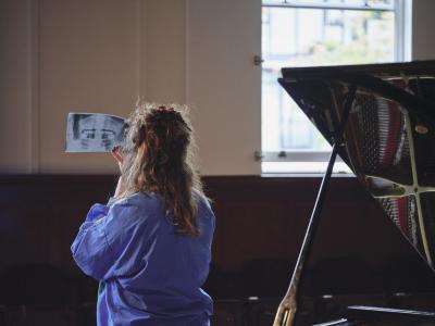 In a daylit Grand Luxe Hall, Eve Risser wears a green, white, purple and yellow windbreaker as they look at an A4 x-ray, facing away from the camera. Beside them stands an open grand piano and their hair is curly in a half up and half down hairstyle.