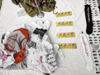 A birds eye view of messy piles of screenprinted clothes, bumper stickers, and knee high socks arranged on a white blanket.