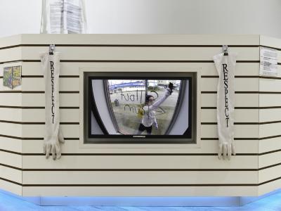 A monitor showing a person behind a window reading sellout scum is embedded in a white slatboard structure. The screen is flanked by two dirty white gloves screenprinted with the word respect.