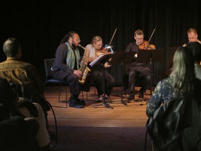 Four musicians, one playing the saxophone, and three the violin, sit next to each other on a wooden stage in spotlight, with a black curtain background. They are framed by seated audience members looking forward.