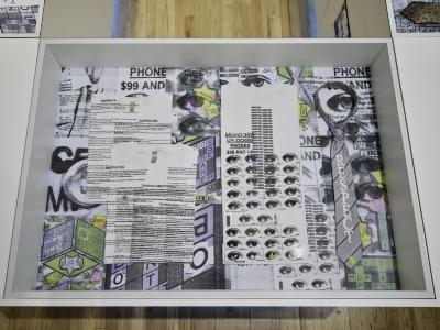 A glass top vitrine displaying white tee shirts screenprinted with dense text and images of eyes, and a necktie reading respect. The inside of the case is lined with enlarged scans of scratch and win lottery tickets.