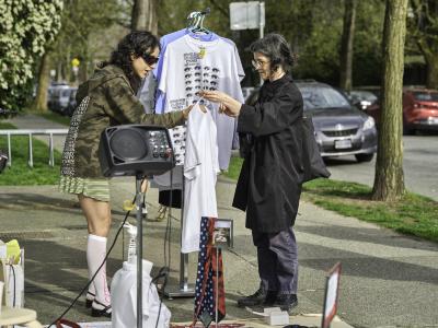 Katayoon Yousefbigloo checks the price tag on a tee shirt held by a customer. They stand in front of a metal clothing rack where a collection of screenprinted tee shirts are on display.