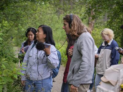Several youth stand on a forest path with sound equipment, staring into a bush. Two wear headphones while others look at their phones in the background. The foreground shows one person with long hair listening and another with dark hair holding up a microphone with a wind muff.