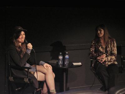 Holly Marie Parnell sits in the foreground to the left, while Christine D’Onofrio, sits to her right in black director chairs. Between them is a black table with two water bottles, and the background is a black wall. Parnell smiles with her gaze off-screen, and a microphone to her face, about to speak. The interviewer holds their microphone at their chest and looks at Parnell with a half-smile.