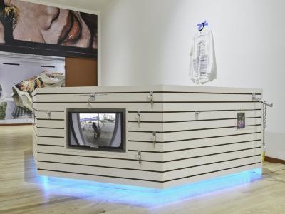 A monitor showing a person at a window is embedded in a multisided kiosk made of white slatboard. Icy blue light emanates from below the structure. A billboard sized photograph of a glamorous Chinese grandmother wraps the wall in the background. 