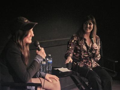Holly Marie Parnell sits on the left side of the foreground out of focus, and the interviewer sits, speaking to her right side in the mid-ground. The interviewer motions to Parnell while they both smile. The background is a black wall, both sit in black director chairs, and a black table sits between them with plastic water bottles and paper notes set down.