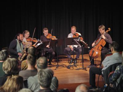 Five musicians sit in a semicircle, one plays the saxophone, three the violin and the fifth a cello, on a wooden stage draped in overhead lighting. Black curtains cover the background and the foreground shows rows of seated audience members.