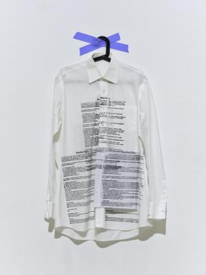 A white dress shirt screen printed with small black text hangs on a black plastic hanger that is adhered to a gallery wall with blue painters tape.