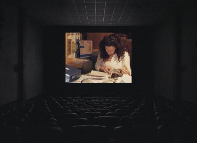 A movie theatre screening shows a person with long brown hair flipping through a full binder of pages. The person’s background is filled with cardboard boxes and other file storage. The cinema is dark, and several people are sitting facing the screen. 