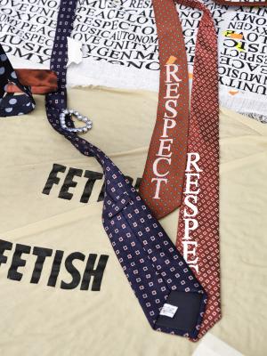 Three ties screenprinted with the word Respect are haphazardly arranged on a beige blanket screenprinted with the word Fetish. One of the ties is ornamented with a rhinestone heart broach. 