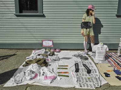 Screenprinted hats, tee shirts and socks are arranged in messy piles on a blanket on the sidewalk alongside a freestanding metal sign with clothing prices. Katayoon Yousefbigloo stands behind the blanket wearing a green dress, sunglasses, and a liquidation world trucker hat. Western Fronts green wood facade provides a backdrop. 