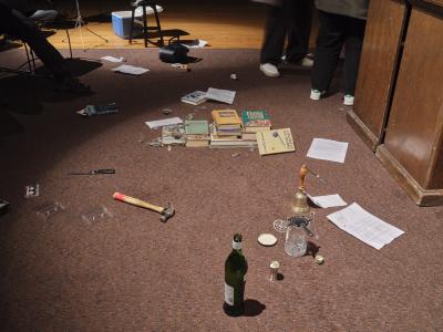 Vintage books, papers, a hammer, a knife, an open bottle of vermouth, a CD player, a cassette, a cocktail kit, and a handbell are all strewn across the spotlit floor of the Grand Luxe Hall.