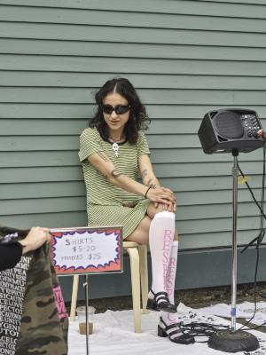 Katayoon Yousefbigloo sits on a stool against Western Fronts green wood facade. She wears black sunglasses, a green striped dress, and knee high socks printed with the word Respect in pink. A metal sign with clothing prices stands on a blanket in front of her.