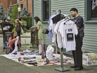 Shoppers browse screenprinted wares at a sidewalk sale, with clothing arranged in piles on blankets on the ground and on a metal rack. Items include ties printed with the word respect and tank tops reading FETISH.