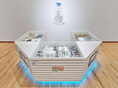 A horseshoe shaped kiosk made from white slatboard displays a variety of screenprinted garments. A white dress shirt is taped to the wall behind. Icy blue light emanates from the bottom of the structure. 