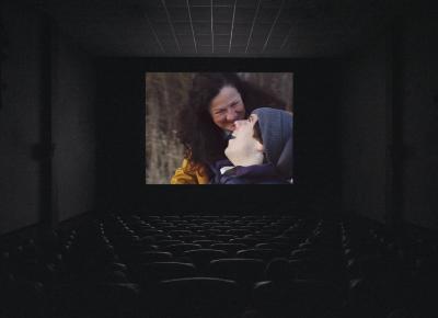 A movie theatre screening shows two people smiling at each other. A person dressed in a grey beanie and blue puffer jacket sits in a wheelchair, smiling in the foreground, their head turned to the left to smile at the person in the mid-ground. This person has long, brown, curly hair and is dressed in a yellow raincoat. The background is a barren winter forest.