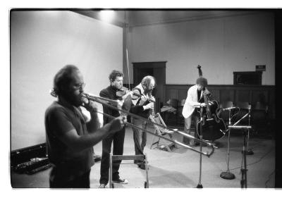 A black-and-white photo of four musicians performing in a line in the Grand Luxe Hall: Gunter Christmann on trombone, David Prentice on violin, Bill Smith on soprano saxophone, and David Lee on cello. 