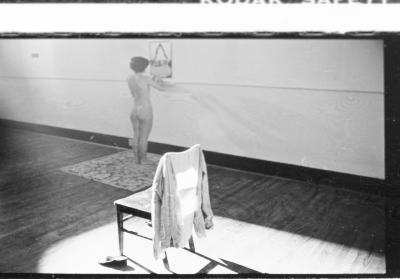 A wooden chair is bathed in sunlight in the gallery. A blouse hangs off its back. Nearby, Jane Ellison stands in the nude on a carpet with her back to the camera as she stares into a mirror that hangs on the wall.