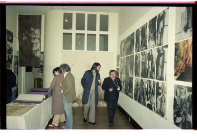 Four people peruse Hermann Nitsch’s exhibition at Western Front. Large-scale photographs are tiled directly onto the gallery wall, and a table with print ephemera is in the middle of the space. 