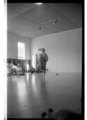 A black-and-white photo showing Jean Erdman balancing on her left foot with her right leg tucked behind and her arms clasped behind her back. Workshop attendees are seated along the wall of EDAM studio.