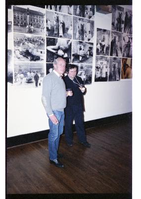 Henry Greenhow and Hermann Nitsch stand in Western Front’s gallery in front of a mural of Nitsch’s photographs. Greenhow wears a grey sweater and blue jeans, and Nitsch is in a dark suit. 