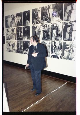 Hermann Nitsch stands in front of an installation of his photographs on the wall of Western Front’s gallery. He wears a blazer and vest over dark-wash pants. His hand is in a cast, and is elevated in a makeshift sling with a string tied around his neck.