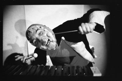 A black-and-white photograph of a television monitor showing Eric Metcalfe playing xylophone. He wears a piranha costume and has a wild smile on his face while holding xylophone mallets in each hand. 