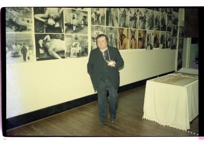 Hermann Nitsch stands in front of an installation of his photographs on the wall of Western Front’s gallery. He wears a blazer and vest over dark-wash pants. His hand is in a cast, and is elevated in a makeshift sling made from a string tied around his neck.