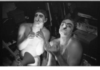 A black-and-white portrait of Martin von Haselberg and Brian Routh posing with their thumbs up. They are both shirtless, and are wearing shower caps and prosthetic noses and chins. Their faces are painted white with black dots to create the effect of stubble. 