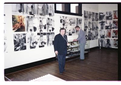 Hermann Nitsch stands in front of an installation of photographs on the wall of Western Front’s gallery. He wears a blazer and vest over dark-wash pants. His hand is in a cast, and is elevated in a makeshift sling with a string tied around his neck.