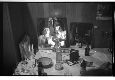 A black-and-white portrait of Martin von Haselberg and Brian Routh in the mirror of their dressing room. Bottles of beer, drinking glasses, scissors, and other objects dot the table where the mirror is positioned.