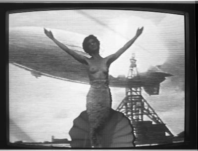 A black-and-white photograph of a television monitor showing Jane Ellison standing with her arms outstretched in front of a projected image of the Hindenburg. Ellison is topless and wears a mermaid tail.