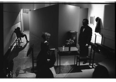 Two performers interact around a table while another lounges in the hall extending their heeled foot into the light. At the table, performers maintain rigid posture while locking eyes.
