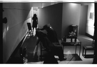 A performer leans back, letting their head drape over the back of a massage chair. Another performer checks on them, holding the sitter's face in their hands, while one more actor looms as a shadow down the hallway. 
