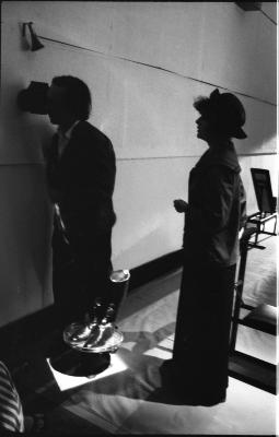 Two performers are separated by a dish holding two upright black boots. One performer rests their eye on a small sculpture mounted to the wall while the other waits, holding something in their hand.
