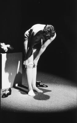 Lit by a spotlight, Jeanette Reinhardt bends over to pull her stockings down over her knees. She is topless and wears only undergarments. Her black patent Mary Janes are on the carpet next to her feet. 