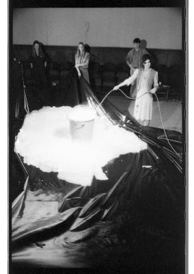 Jane Ellison snakes a tube into a plastic garbage bin that is overflowing with bubbles. Three people stand on the periphery of a tarp that catches the spill. 