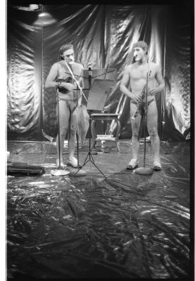 Brian Routh plays ukulele and Martin von Hasselberg holds a clarinet. They stand at microphones and are dressed in swim caps and jockstraps. Tarp covers the floor and back wall of the Grand Luxe Hall.